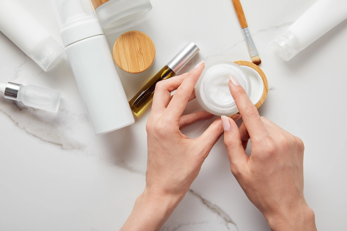 A pair of hands opening a jar of age management cream, surrounded by various other anti aging skin care and aging skincare products on a marble surface.